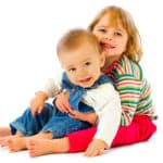 photo of a baby in blue overalls on the lap of a young girl with both smiling