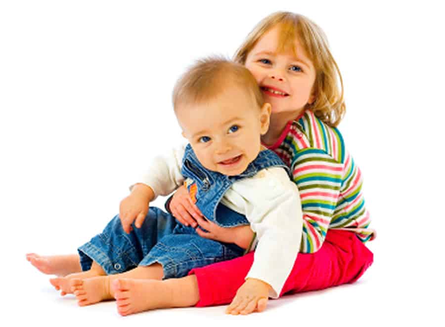 photo of a baby in blue overalls on the lap of a young girl with both smiling