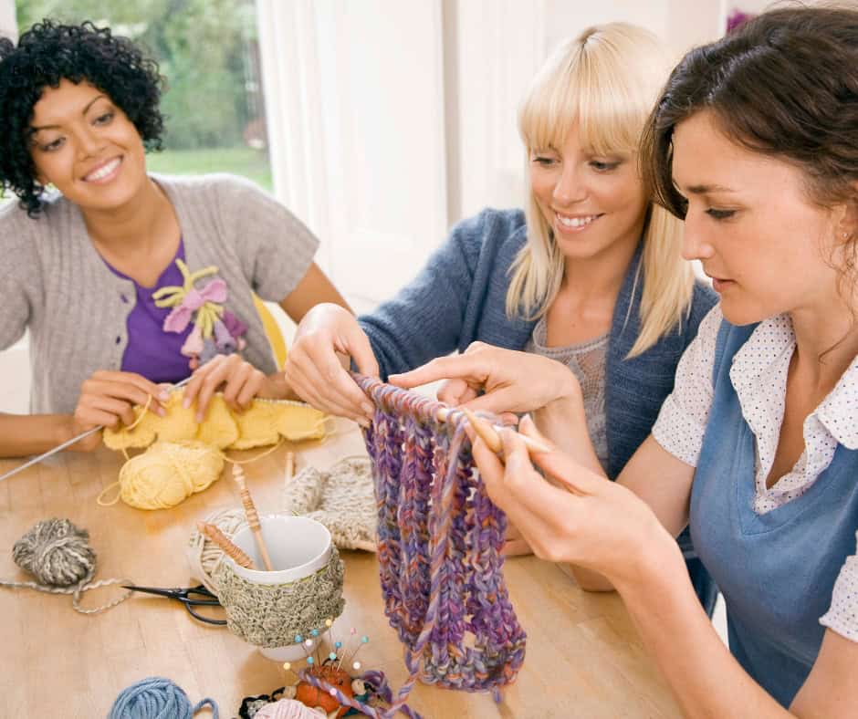 A group of three women knitting together. One woman has tan skin and dark curly hair, one has pale skin and long blonde hair, and the last has pale skin and wavy brown hair.