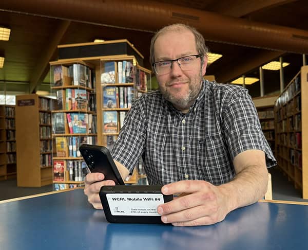 image of a man holding a small black device in one hand and a black case in the other seated at a table in the library.