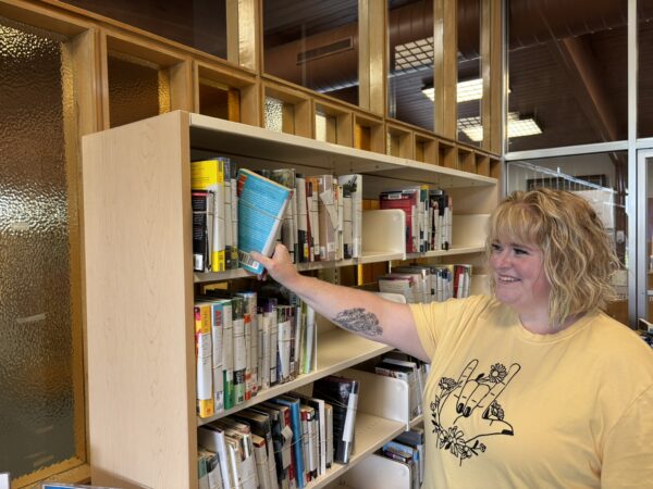 image of a woman with blonde hair and yellow shirt reaching for books on a shelf.