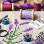 A cross-stitch of a lavender stalk. It is surrounded by spools of thread, scissors, needles, and a real stalk of lavender.