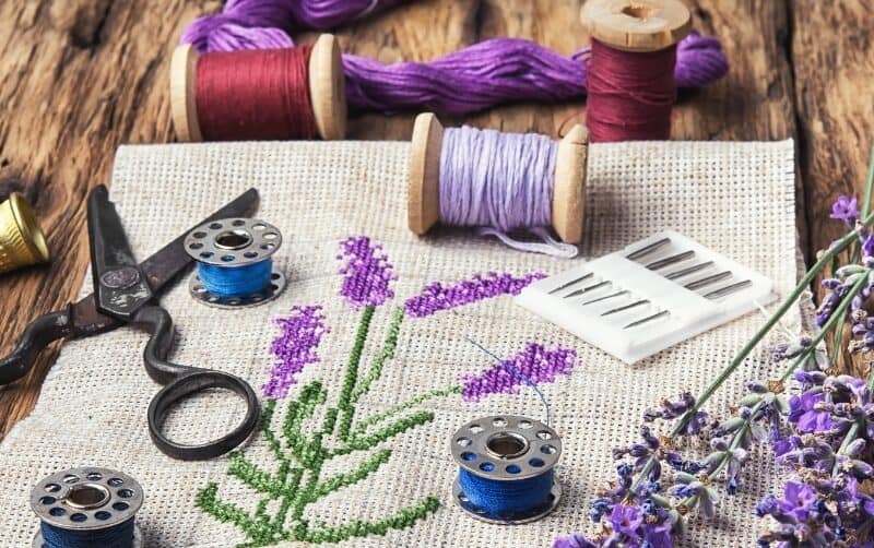A cross-stitch of a lavender stalk. It is surrounded by spools of thread, scissors, needles, and a real stalk of lavender.