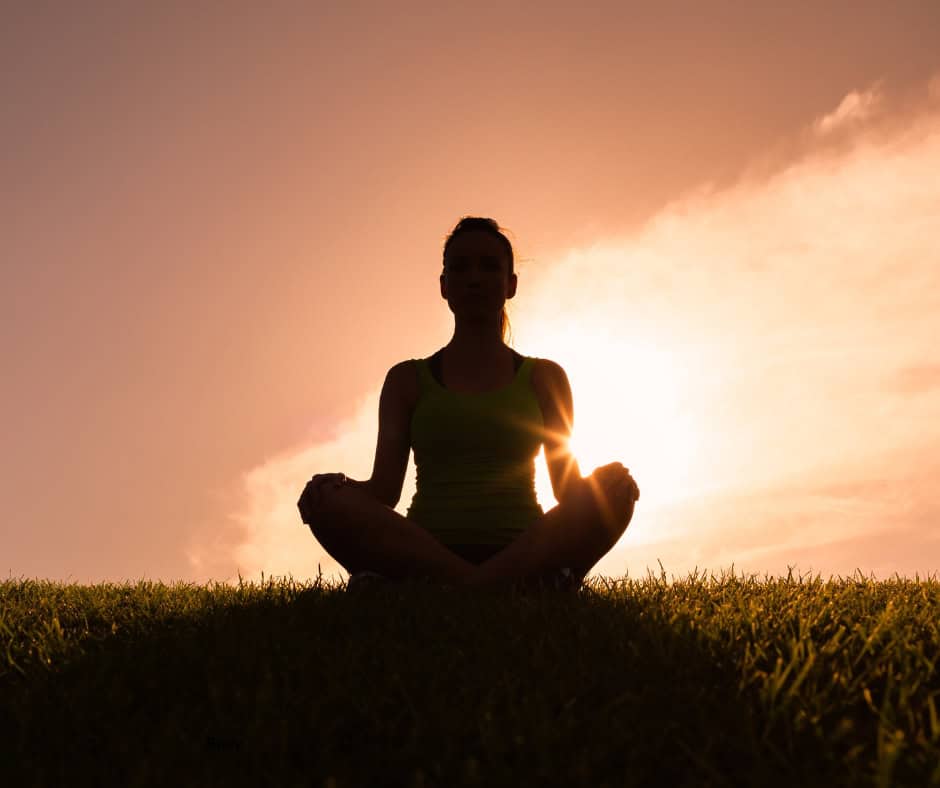 The silhouette of a woman sitting with her legs crossed, meditating on a grassy plain. Behind her is an orange sunset.