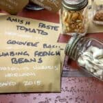 Seeds preserved in brown paper packets and in jars. The packets are named, Climbing French Beans, Genovese Basil, and Amish Paste Tomato.
