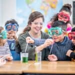 A group of children with different ethnicities. They surround a young woman who shows them how to do a science experiment with beakers. They all have protective eyewear on.