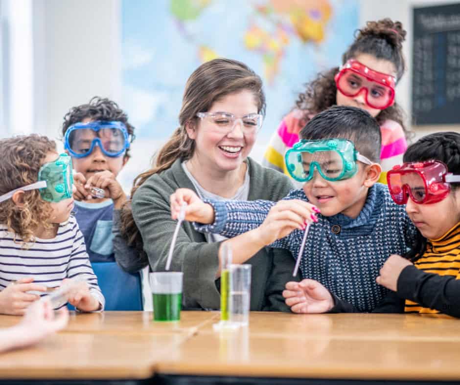 A group of children with different ethnicities. They surround a young woman who shows them how to do a science experiment with beakers. They all have protective eyewear on.