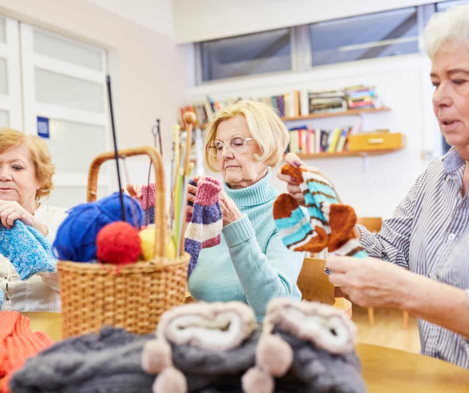 Three older white women sit at a table. They are admiring the socks they knitted.