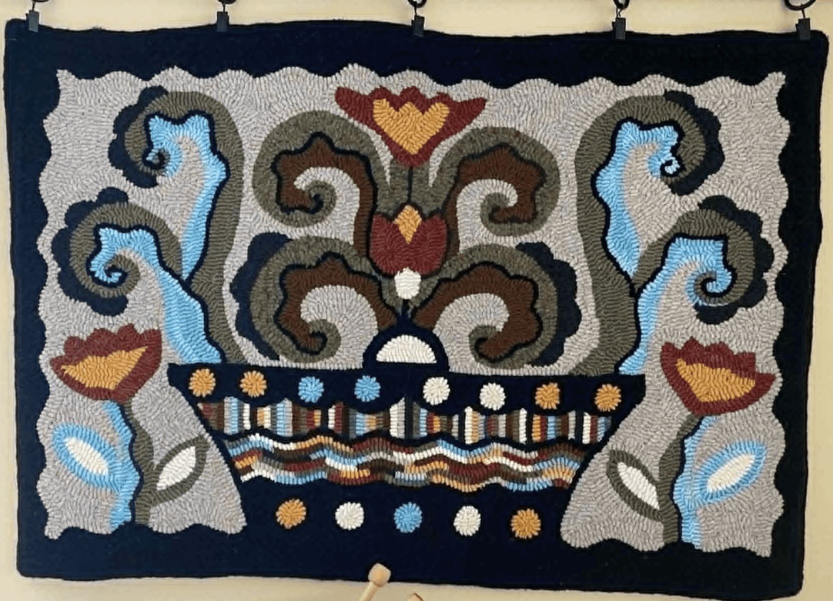 A tapestry that has been rug hooked. It is an interpretation of a pot with flowers and vines growing out of it.
