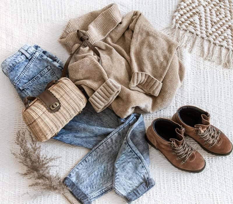 Women's fall clothing folded on a bed. The outfit includes, jeans, boots, a knitted sweater, and a small wicker handbag.