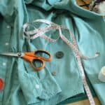 A blue dress shirt that is prepared for mending. There is a needle and thread in the sleeve with two loose buttons next to it. Someone has also placed a measuring tape, thread, and scissors nearby.