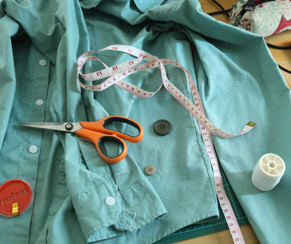 A blue dress shirt that is prepared for mending. There is a needle and thread in the sleeve with two loose buttons next to it. Someone has also placed a measuring tape, thread, and scissors nearby.