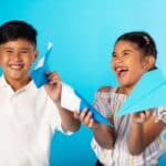 A boy and girl smiling and holding paper airplanes in their hands. The background is blue and so are the airplanes.