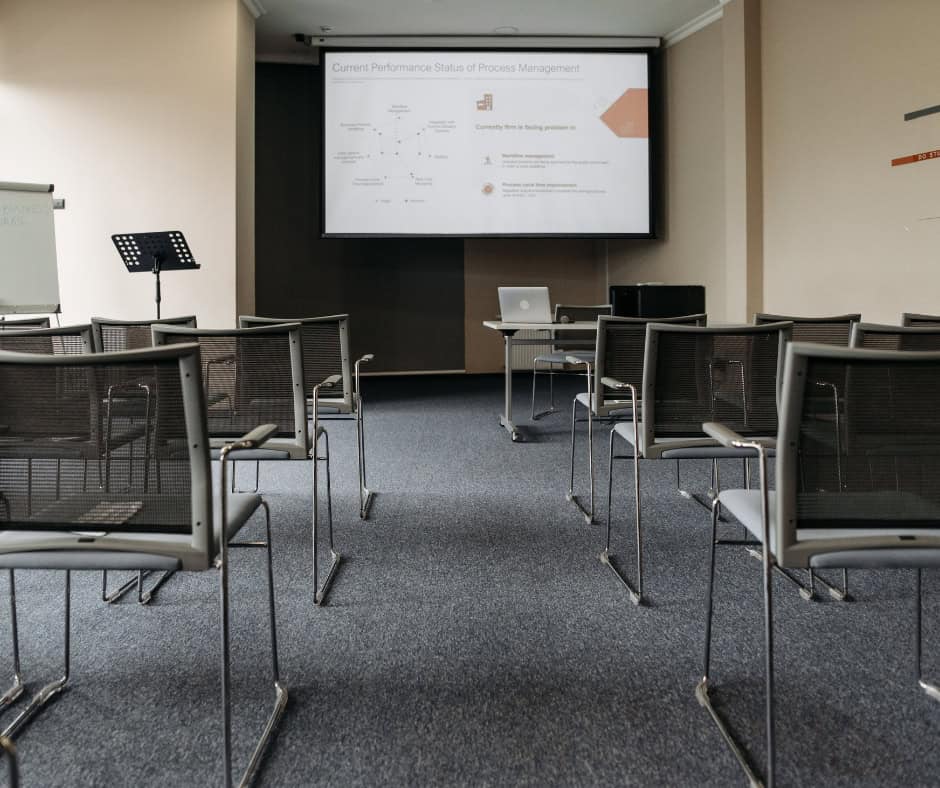 A room lined with metal chairs. At the back of the room is a projector screen with a presentation being shown.