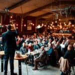 A large group of people seated at tables playing a trivia game. One man stands in the forefront and asks them questions.
