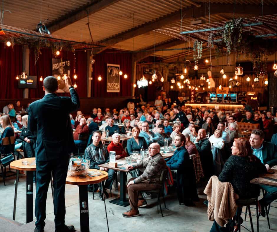 A large group of people seated at tables playing a trivia game. One man stands in the forefront and asks them questions.